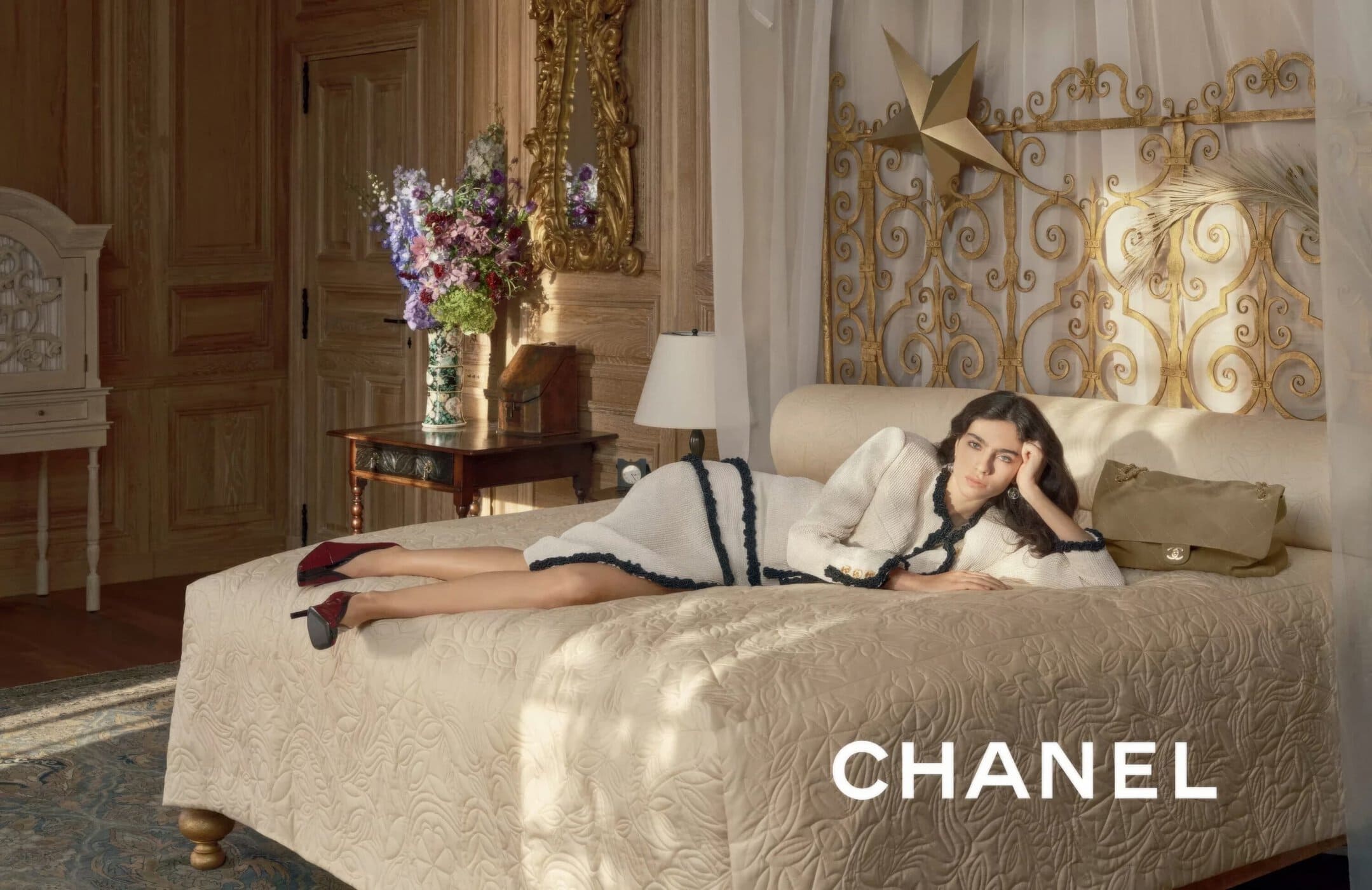 Model lying on a cream quilted bed wearing a white and dark blue trim tweed jacket and skirt set in a classic room