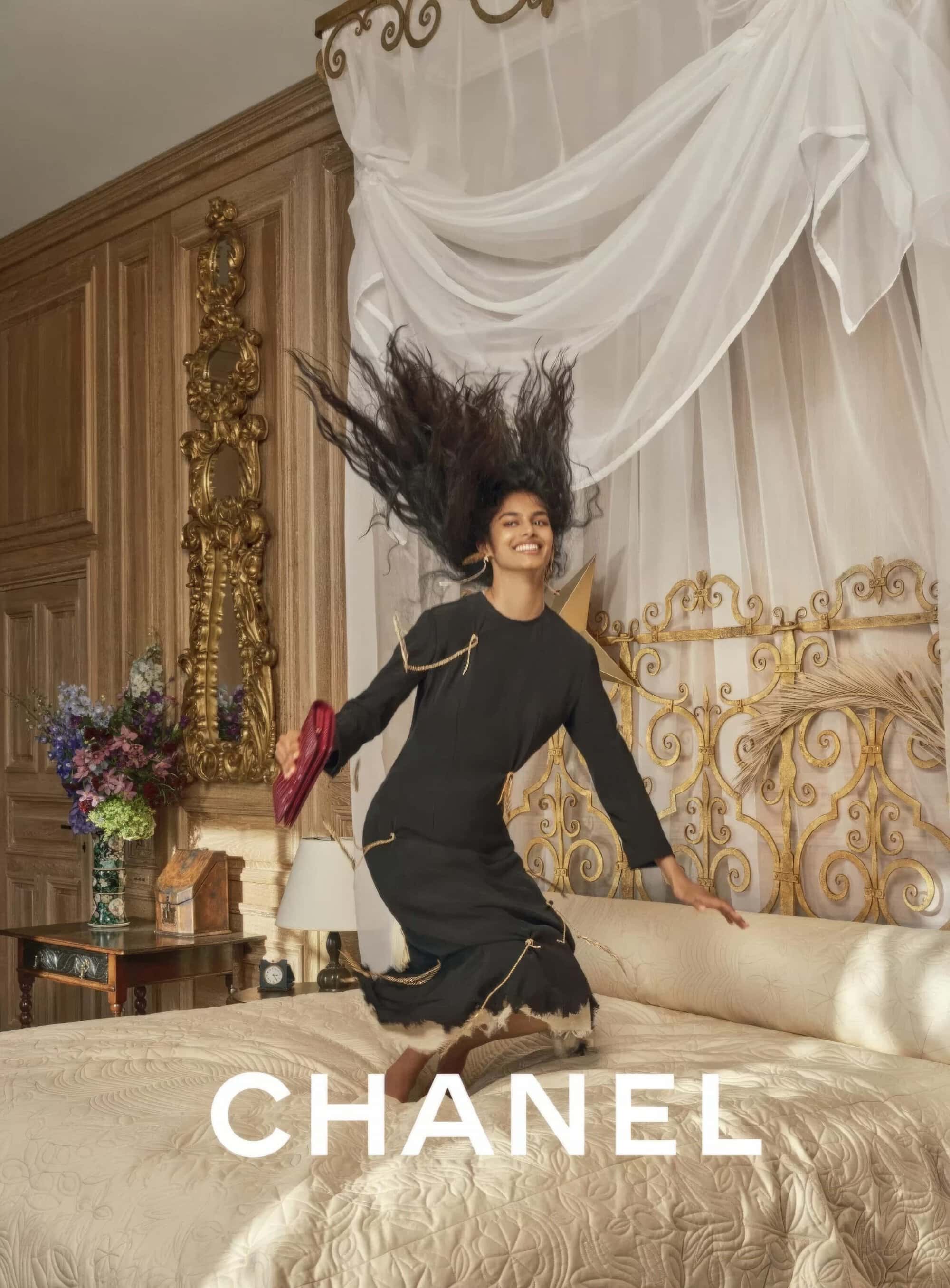 Smiling model jumping on a luxury bed in a black dress holding a red clutch with draped white canopy overhead.