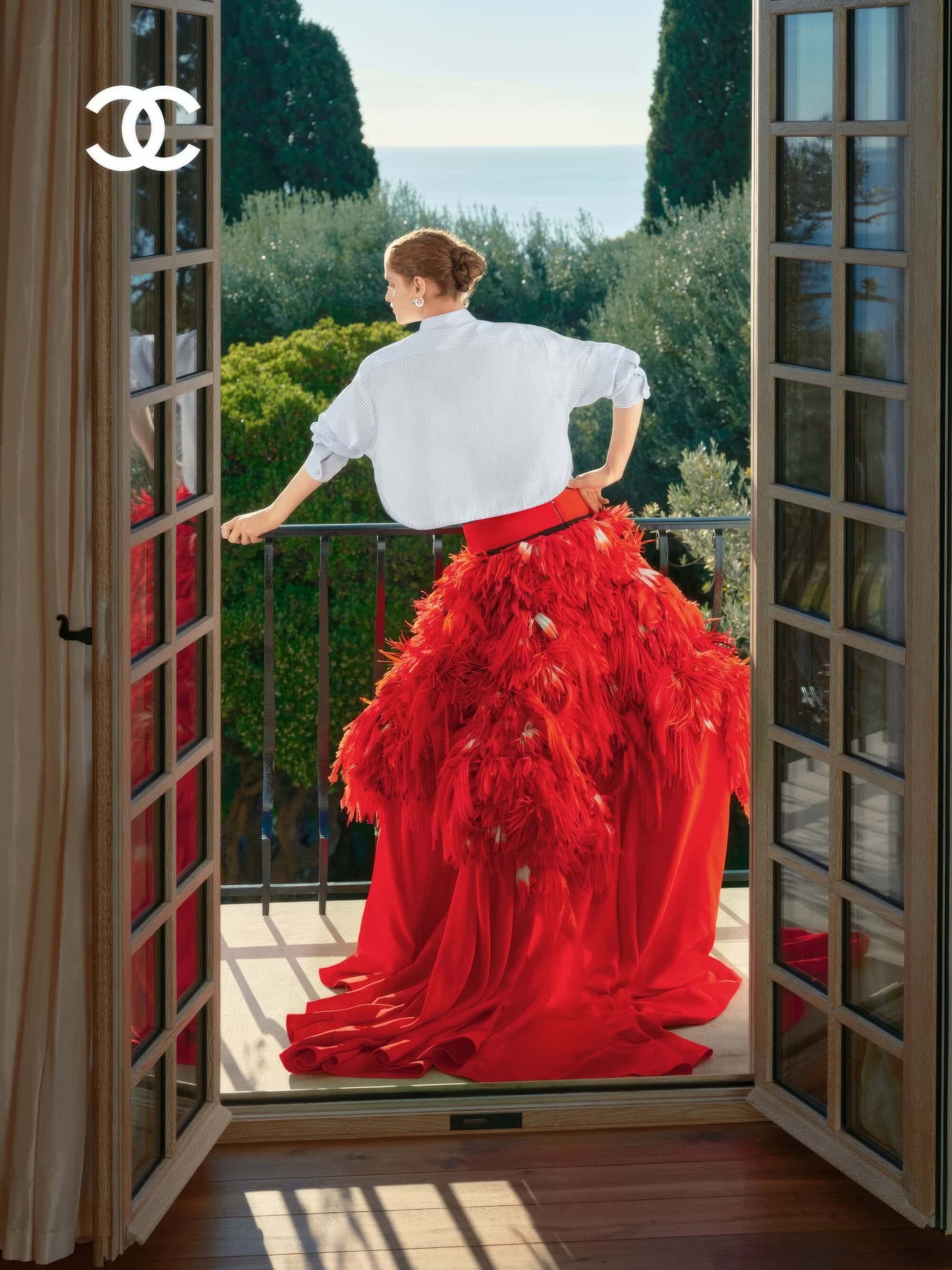 Model wearing a white shirt and voluminous red skirt on a balcony overlooking the sea, framed by open French doors.