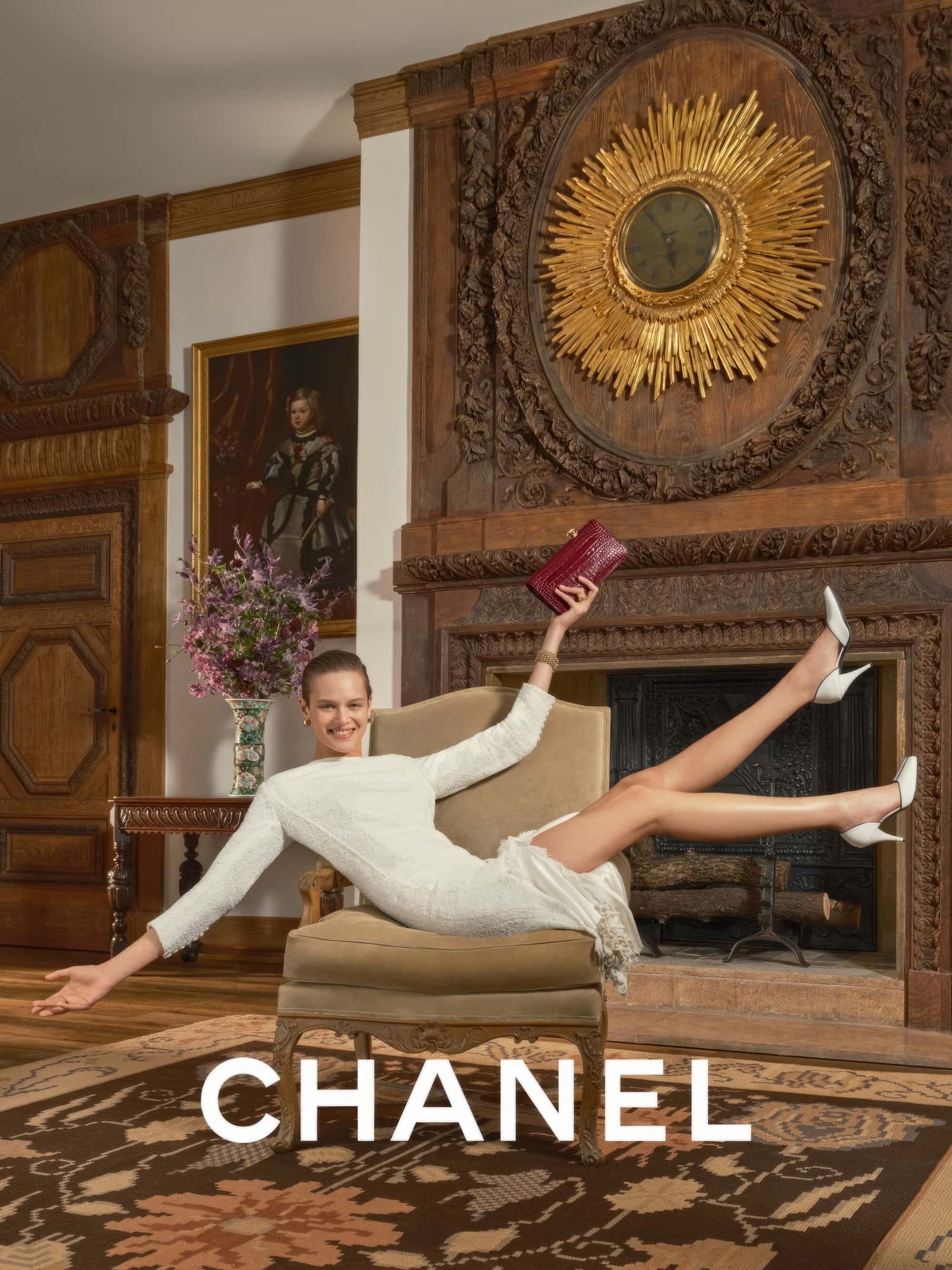 Model wearing a white dress holding a red clutch in an ornate room with a fireplace
