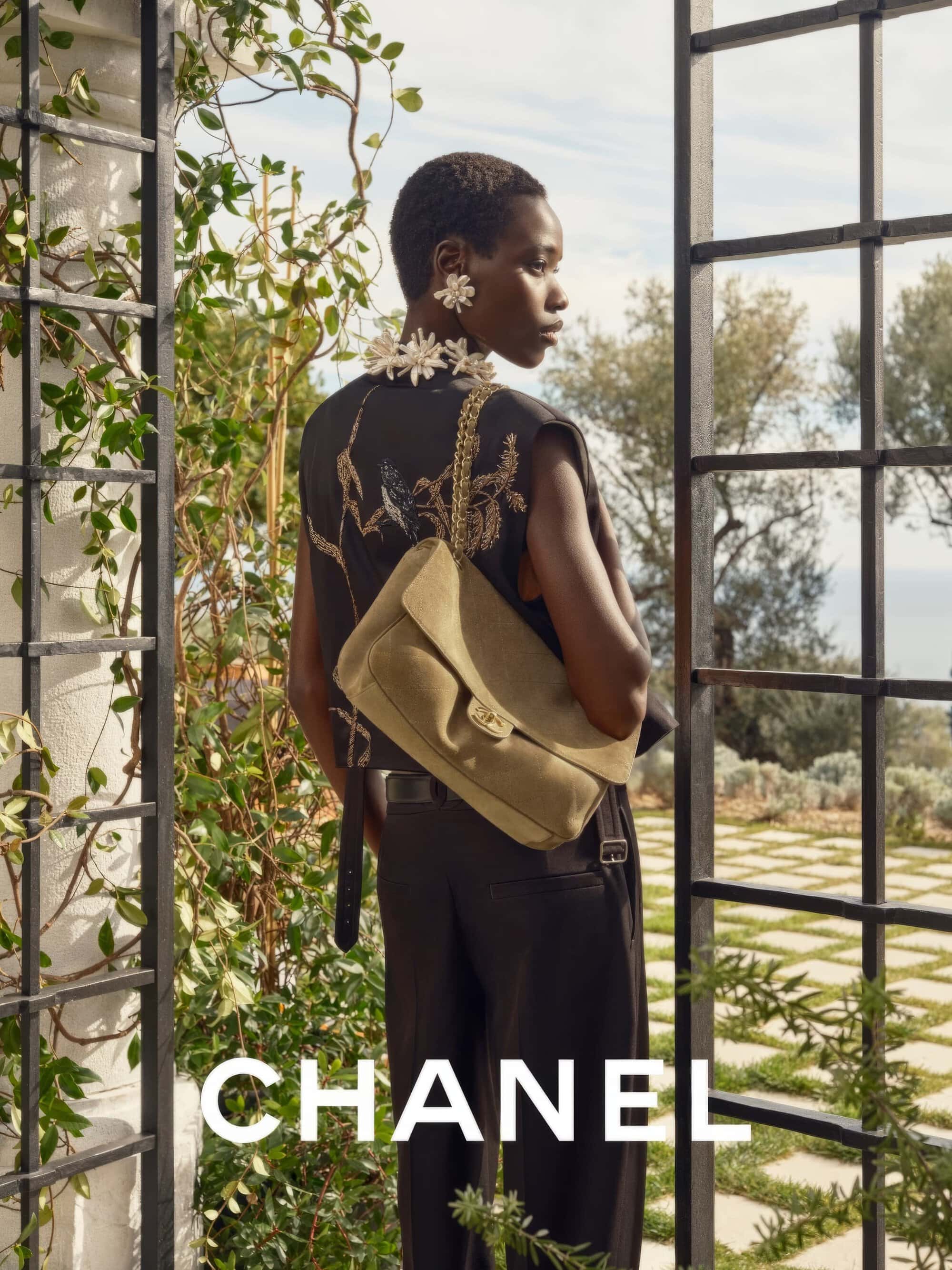Model in dark sleeveless embroidered top wearing a tan suede shoulder bag outdoors near a trellis.