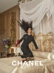 Smiling model jumping on a luxury bed in a black dress holding a red clutch with draped white canopy overhead.