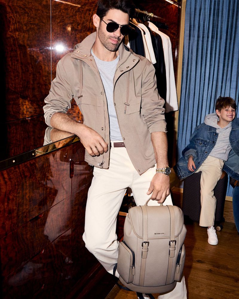 Stylish adult man with backpack poses next to boy. Man in casual clothing and sunglasses, boy in jean jacket. Indoor setting.