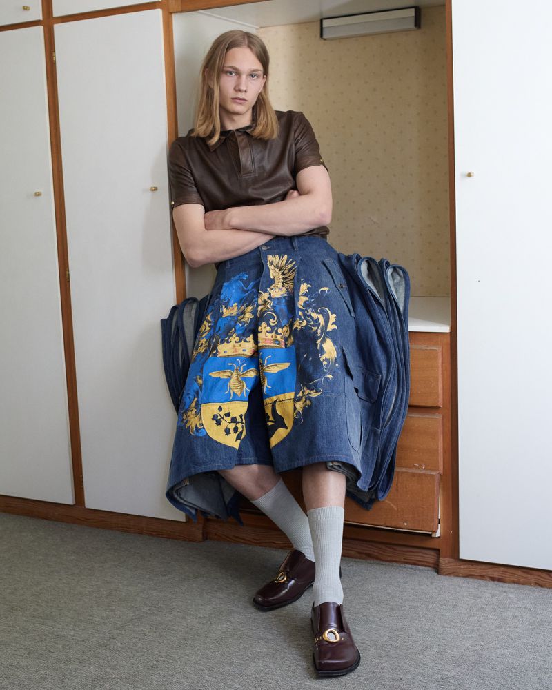 A young male model with long blonde hair leaning against a set of lockers wearing a brown leather top and printed blue denim shorts