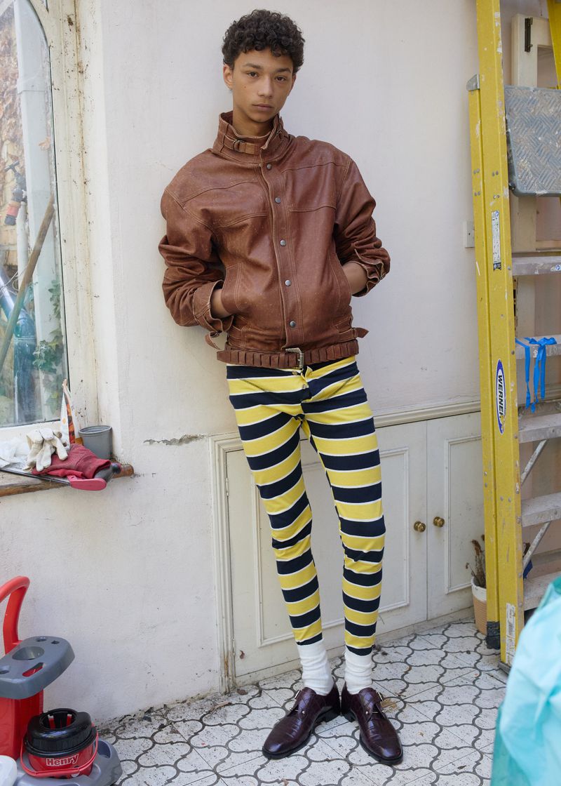 Model leaning against a wall wearing a brown leather jacket and striped yellow trousers.