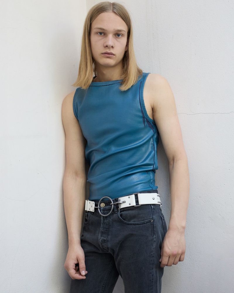 Young male model with long blonde hair wearing a blue sleeveless top and white O-ring belt standing against a white wall.