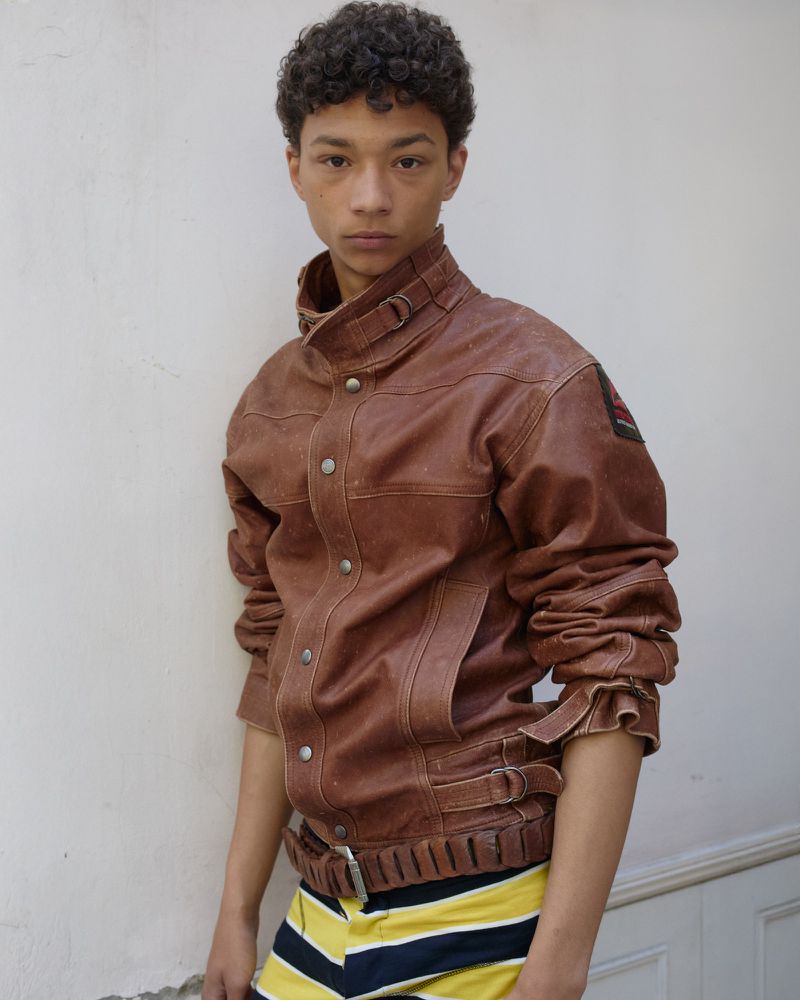 Young male model with dark curly hair wearing a brown leather jacket against a plain white wall.