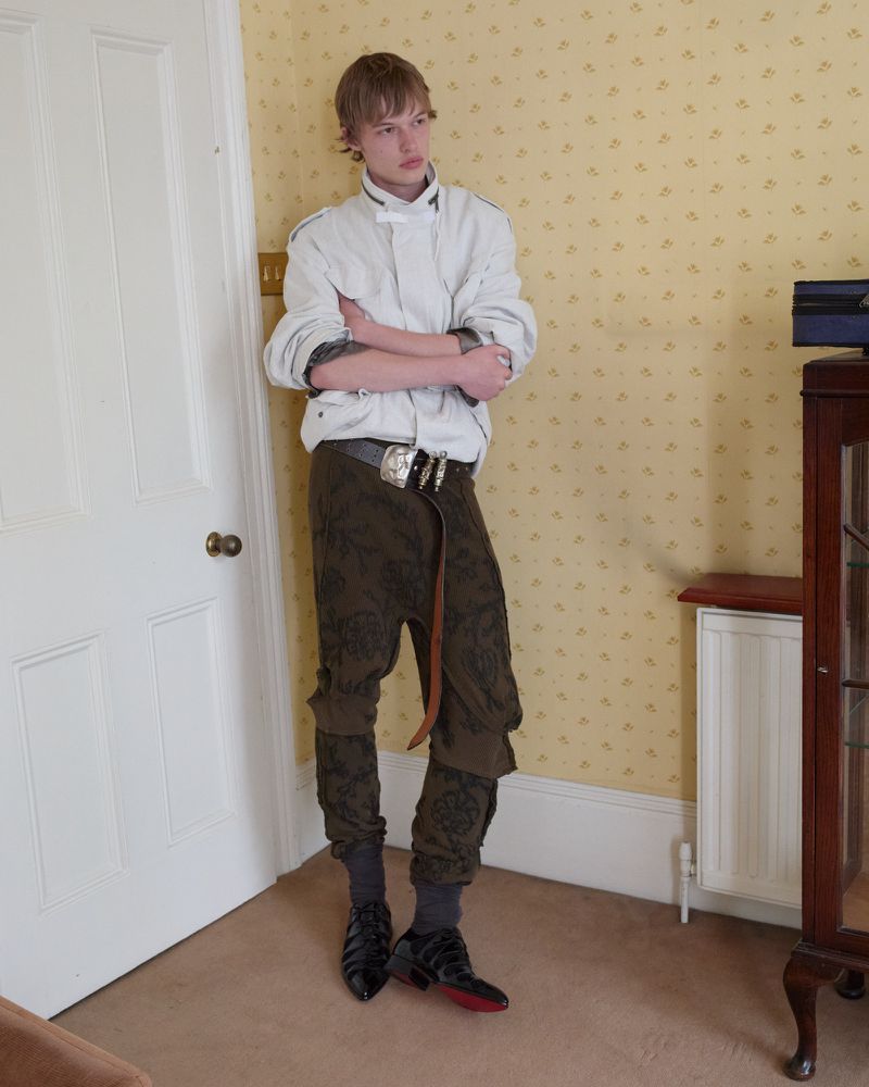 Young male model standing indoors in a white jacket and patterned trousers against patterned wallpaper.