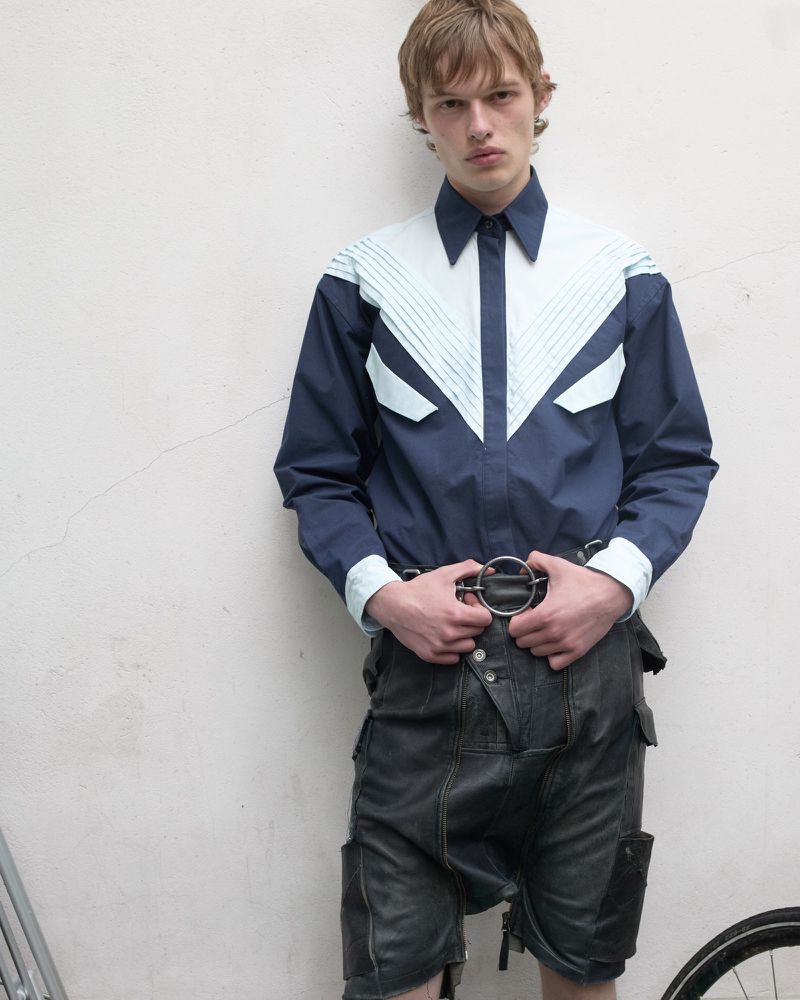 Model wearing a navy and light blue paneled shirt and black shorts against a plain white wall.