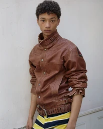 Young male model with dark curly hair wearing a brown leather jacket against a plain white wall.