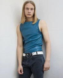 Young male model with long blonde hair wearing a blue sleeveless top and white O-ring belt standing against a white wall.
