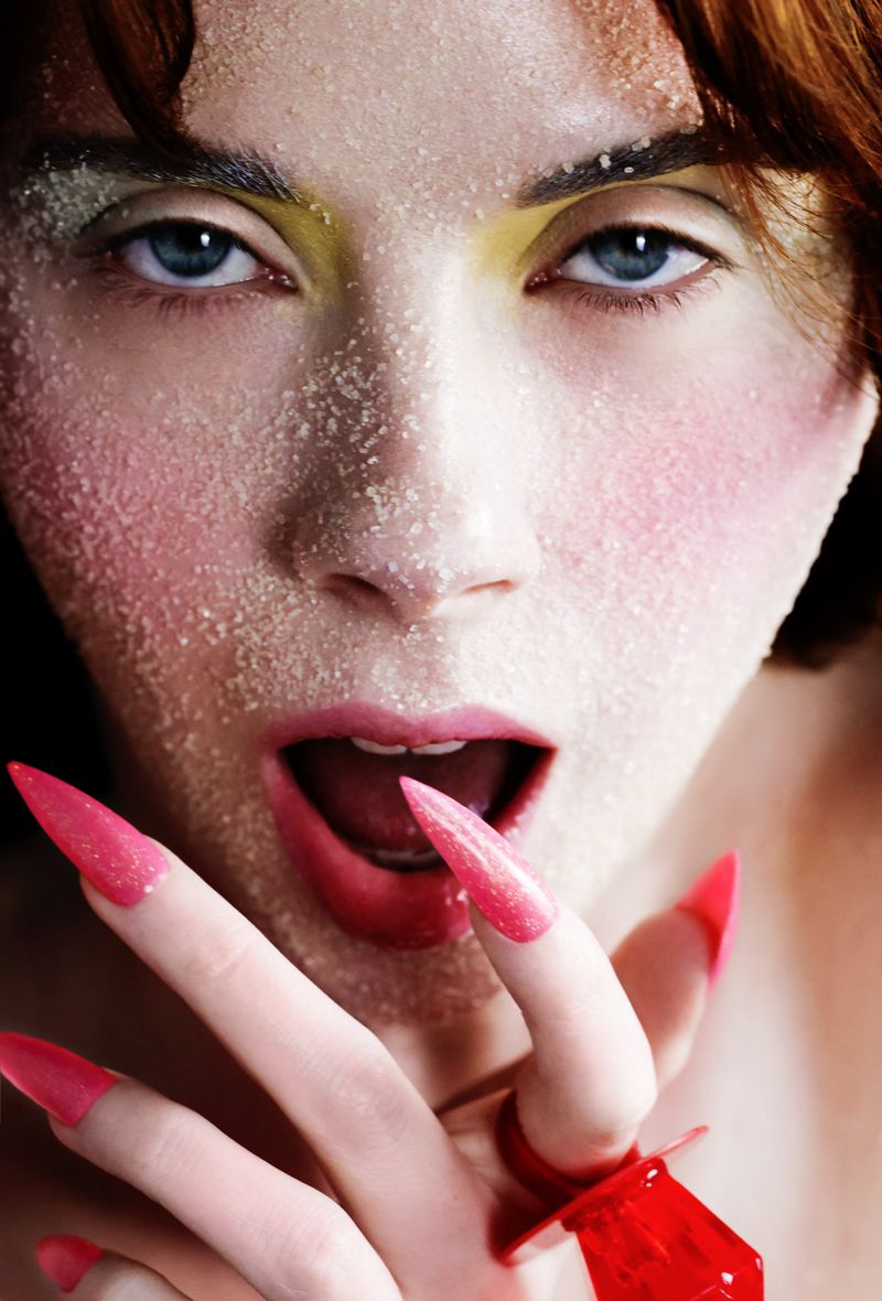 Close-up portrait of a woman with sugar on her face, pink lipstick, and long pointy pink fingernails.