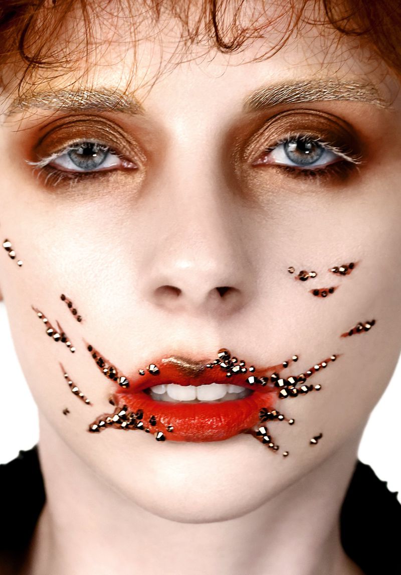 Close-up portrait of a model with metallic beads on cheeks and lips, golden eyebrows, and vibrant red lipstick against a white background.