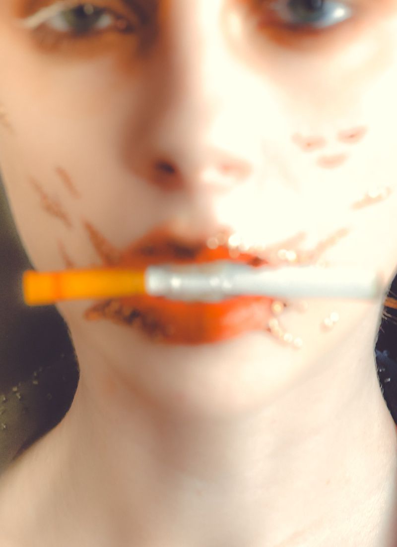 Close-up portrait of a person holding a cigarette between their lips with smeared red lipstick and marks on their face