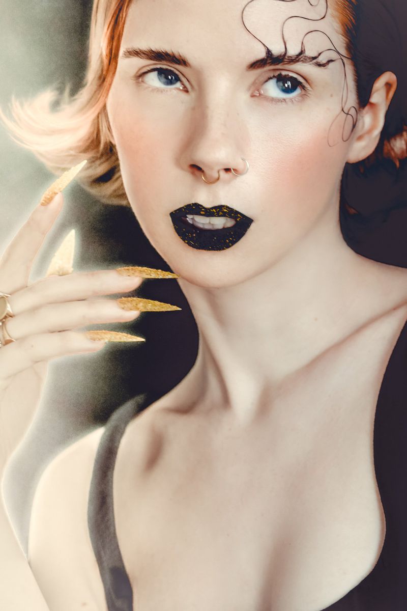 Close-up portrait of a woman with dark lipstick, gold sparkly long nails, and gelled hair swirls on her forehead by Brenda Nasr.