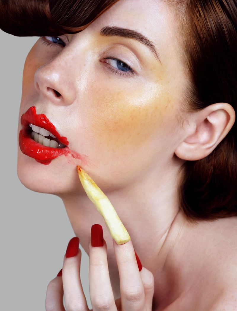 Close-up portrait of a woman with smudged red lipstick holding a french fry against her cheek, photographed by Brenda Nasr.