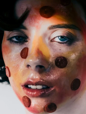 Close-up portrait of a woman with painted face and small pepperoni-like food items adhered to her skin