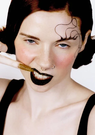 Close-up portrait of a woman with dark lipstick touching her tooth with a textured golden fingernail against a plain white background.