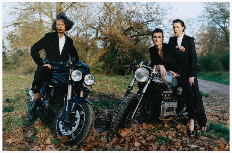Three models in black blazers posing on motorcycles in a wooded area with fallen leaves, photographed by Estelle Hanania.
