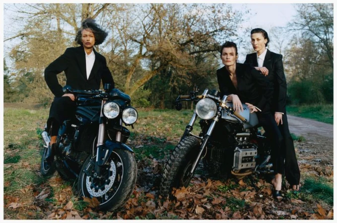 Three models in black blazers posing on motorcycles in a wooded area with fallen leaves, photographed by Estelle Hanania.
