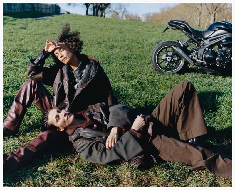 Two women wearing dark jackets and trousers seated on a grassy hillside with a parked motorcycle