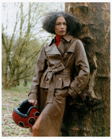 Model leaning against a tree wearing a brown leather coat and holding a red helmet in a forest