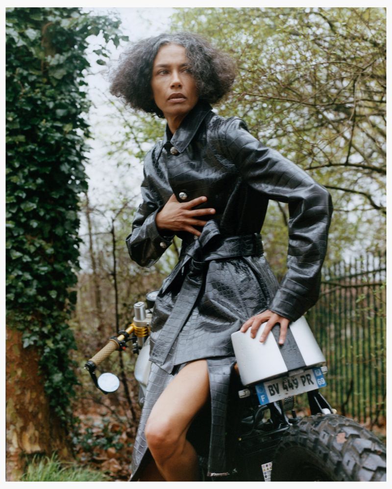 Model with dark textured hair wearing a black glossy belted coat sitting on a motorcycle outdoors.