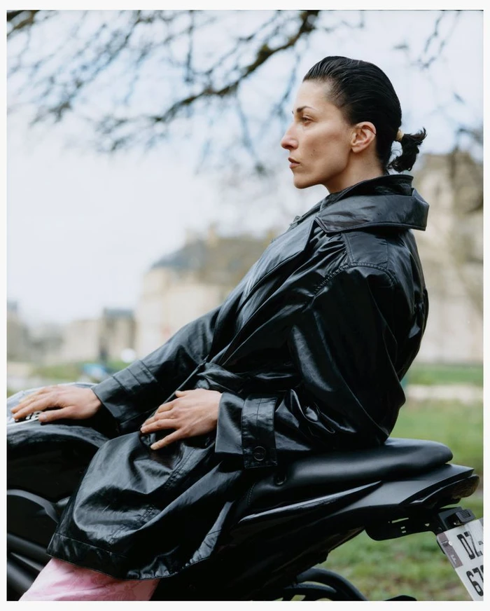 Model in a black leather trench coat leaning on a motorcycle outdoors with blurred architecture and trees in the background