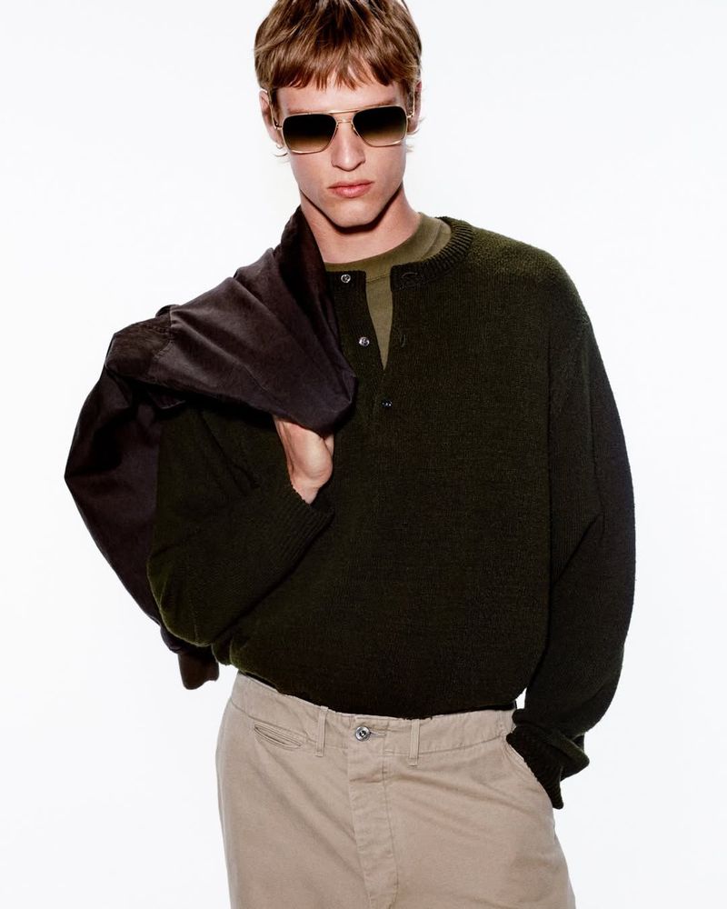 Male model wearing tinted sunglasses, dark green cardigan, and beige trousers with a jacket draped over his shoulder.