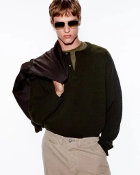Male model wearing tinted sunglasses, dark green cardigan, and beige trousers with a jacket draped over his shoulder.