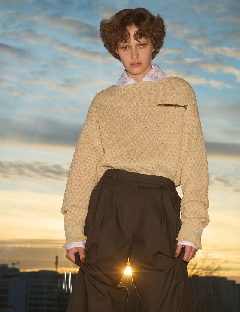 Model wearing a textured camel sweater and dark trousers against a sunset sky backdrop for System Collections.