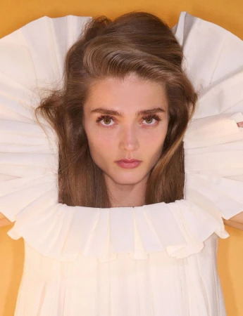 Portrait of a model wearing a pleated white collar framing her face against a warm yellow background.