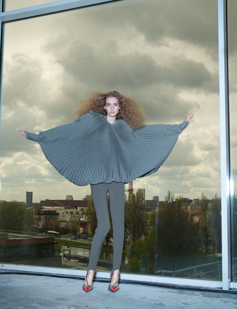 Model wearing a grey pleated cape top and leggings standing in front of a large window with city skyline view