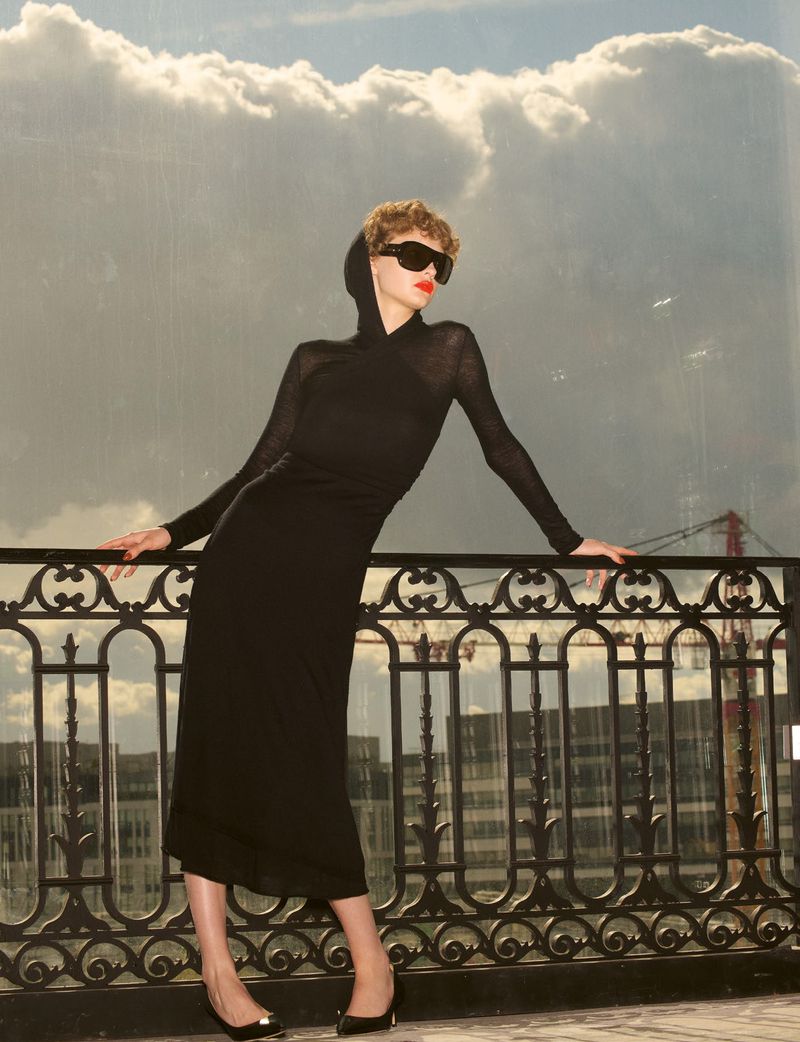 Model in a black hooded dress and sunglasses leaning against an ornate balcony railing with a cloudy sky background.