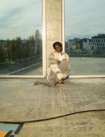 Model seated on concrete floor wearing patterned fur jacket against large windows for System Collections.