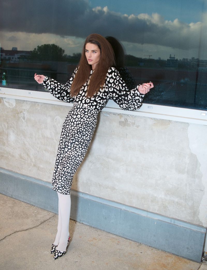 Model in a black and white patterned dress leaning against a wall outdoors under a cloudy sky.