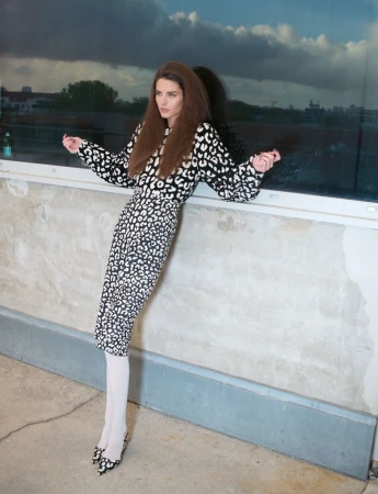 Model in a black and white patterned dress leaning against a wall outdoors under a cloudy sky.