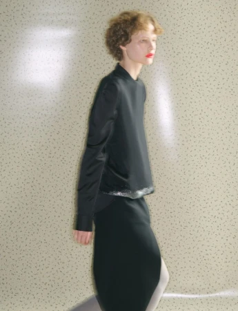 Model with curly hair and red lipstick wearing a black top and skirt against a patterned backdrop for System Collections.