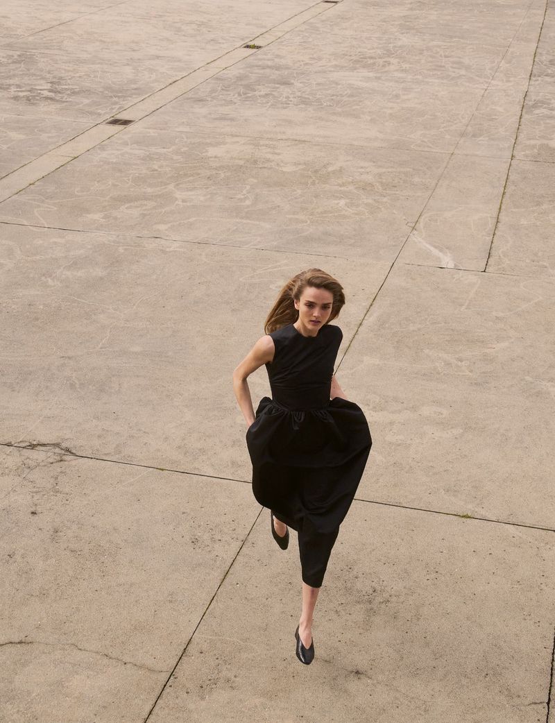 Model in a black sleeveless dress running across a vast concrete plaza from a high-angle view