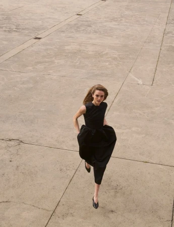 Model in a black sleeveless dress running across a vast concrete plaza from a high-angle view