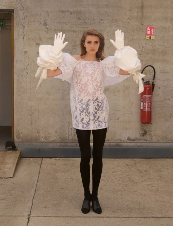 Model in white lace dress with puffed sleeves and white gloves posing against a concrete wall for System Collections.