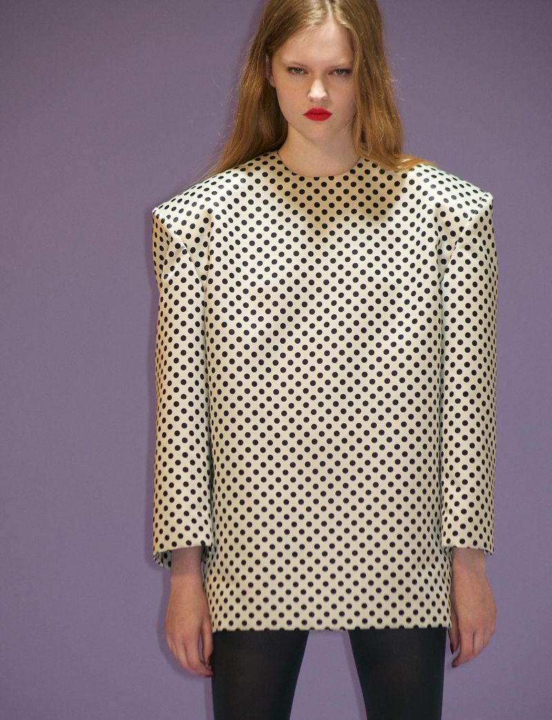 Model wearing a structured polka dot top with prominent shoulders against a plain purple backdrop