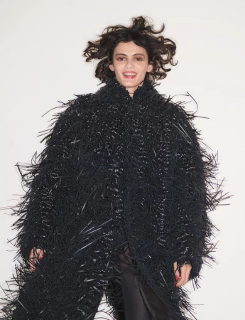 Model wearing a textured black fringed coat with tousled dark hair against a plain white studio background.