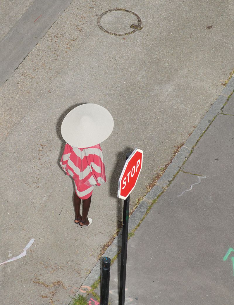 High-angle shot of a person wearing a pink and white striped dress and large white hat standing next to a stop sign on pavement.