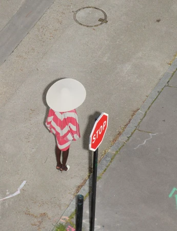 High-angle shot of a person wearing a pink and white striped dress and large white hat standing next to a stop sign on pavement.