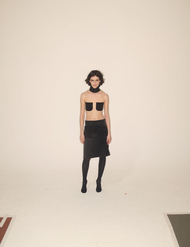 Model wearing dark clothing and a neckpiece standing against a plain white studio backdrop