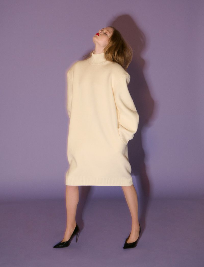 Model wearing a cream turtleneck dress and black heels against a purple background