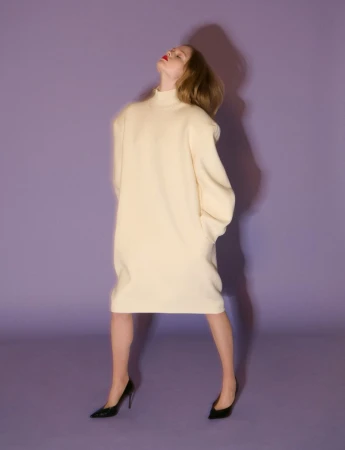 Model wearing a cream turtleneck dress and black heels against a purple background