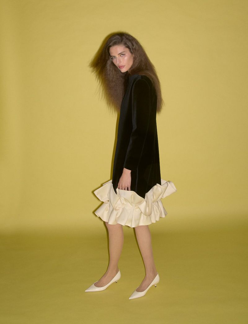 Model in black dress with cream ruffled hem and white heels against yellow background for System Collections