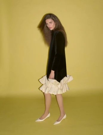 Model in black dress with cream ruffled hem and white heels against yellow background for System Collections